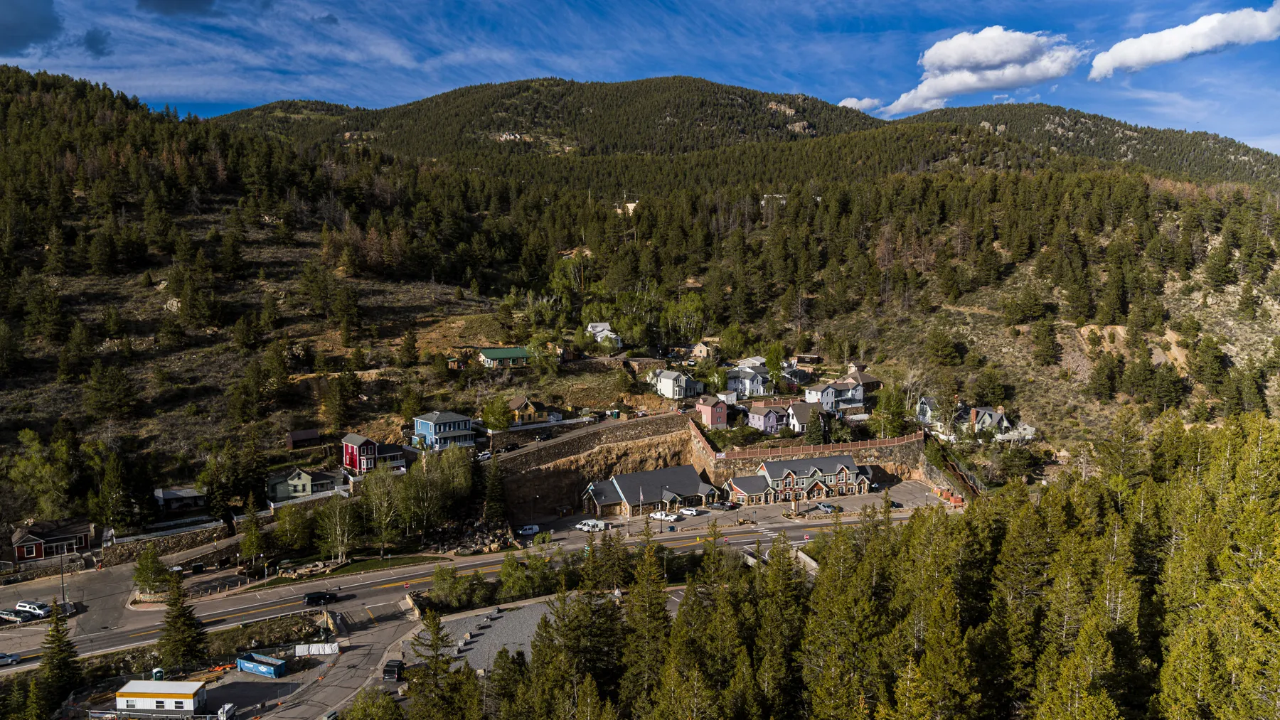 Aerial view of Black Hawk, Colorado showing small mountain town with scattered buildings and homes built on hillsides. The historic mining settlement sits in a valley surrounded by dense pine forests and Rocky Mountain peaks.